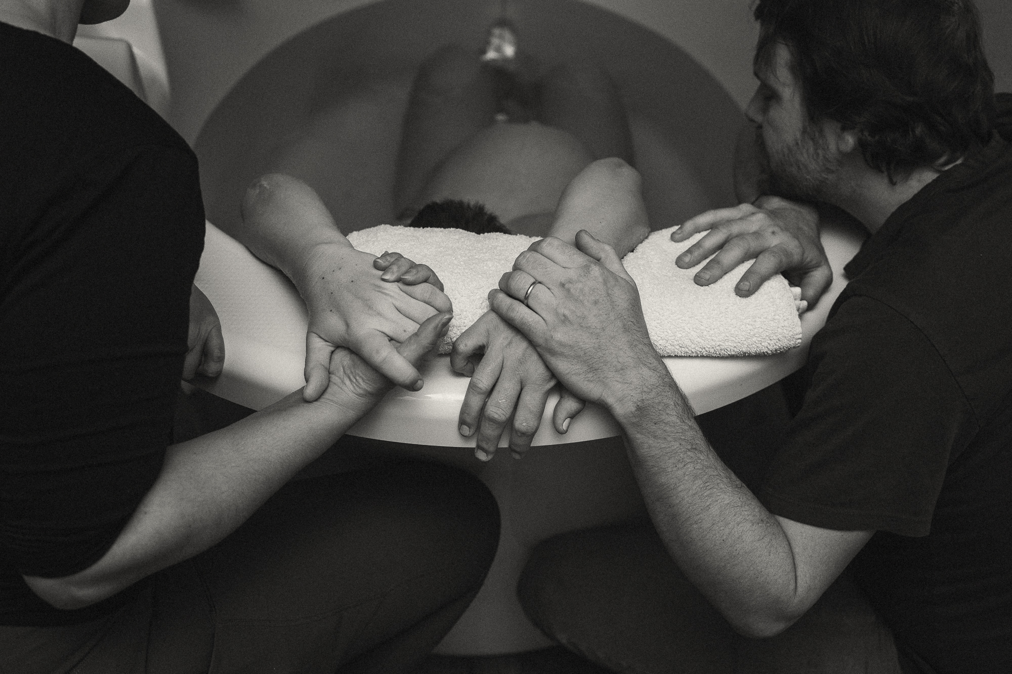 black and white image of a laboring person in a tub, viewed from above with just their belly, legs and hands visible; the hands of support people are visible grasping each of the pregnant person's hands as they support her from the edges of the rounded birthing tub.